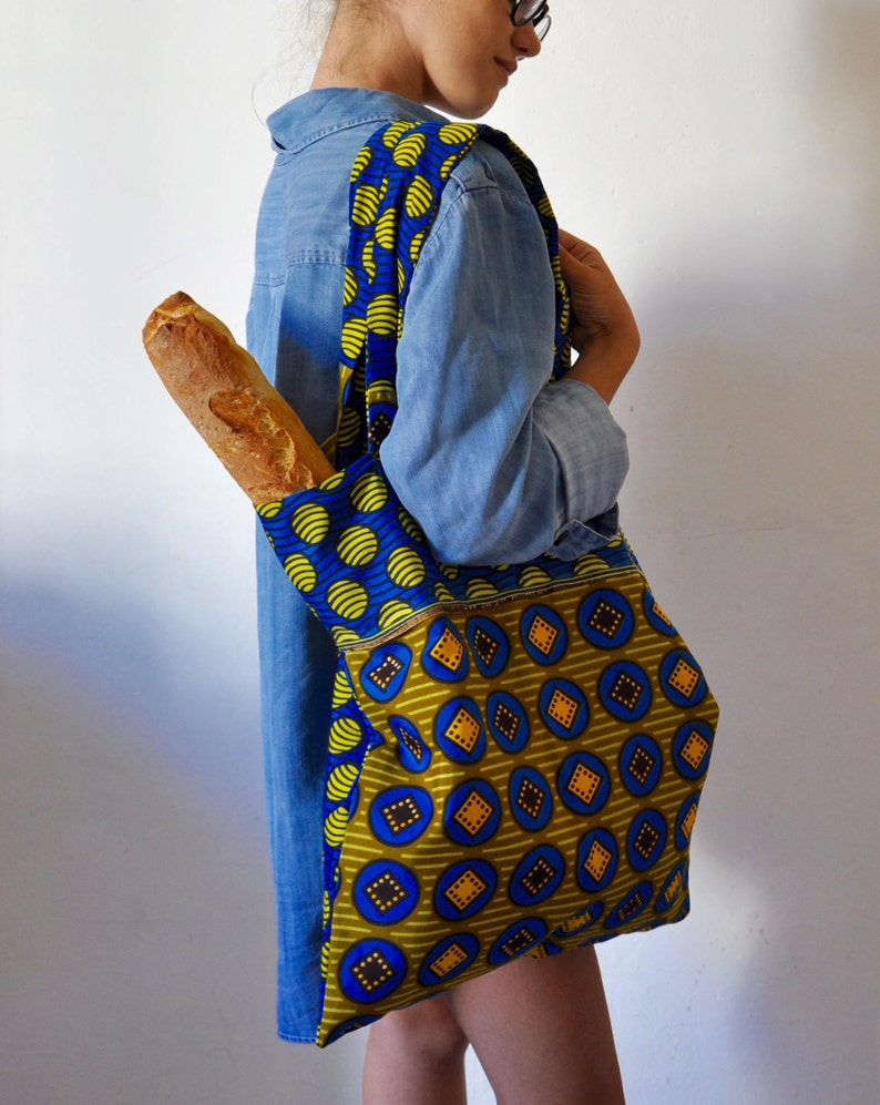 May include: A woman is carrying a blue and yellow patterned tote bag with a baguette inside. The bag has a large pocket on the front with a different pattern.