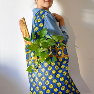 May include: A woman is carrying a blue and yellow tote bag with a plant and a baguette inside. The bag has a geometric pattern on the top and a polka dot pattern on the bottom.