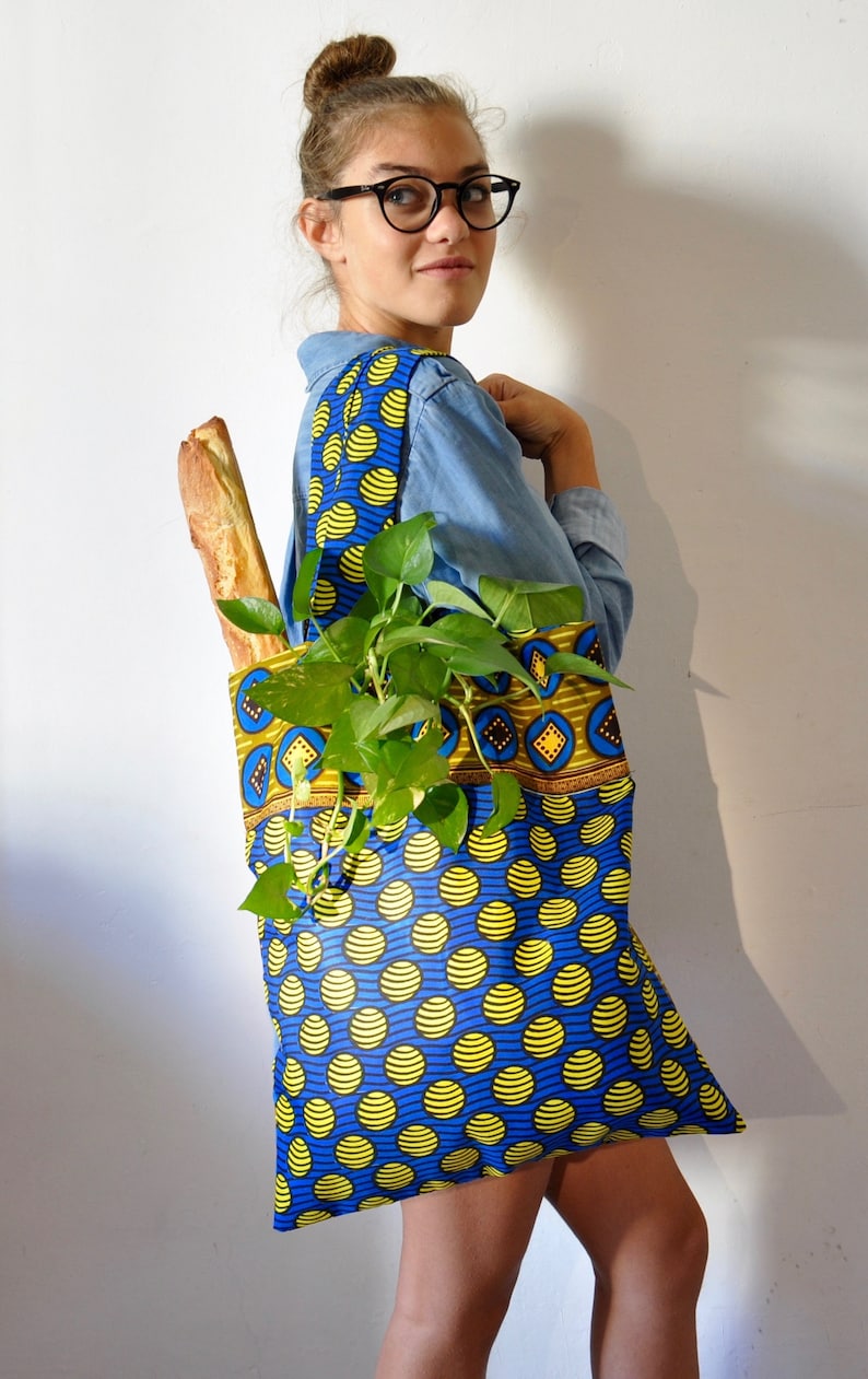 May include: A woman is carrying a blue and yellow tote bag with a plant and a baguette inside. The bag has a geometric pattern on the top and a polka dot pattern on the bottom.