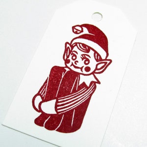May include: A white gift tag featuring a red illustration of a Christmas elf. The elf is depicted in a red hat and outfit. The tag has a hole at the top for hanging, suitable for festive occasions.