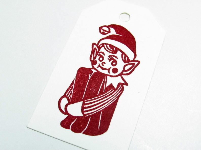 May include: A white gift tag featuring a red illustration of a Christmas elf. The elf is depicted in a red hat and outfit. The tag has a hole at the top for hanging, suitable for festive occasions.
