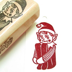 May include: A wooden stamp featuring a carved image of a smiling elf in a hat and collared shirt. Beside it is a red ink impression of the elf. The stamp has the text "witchcraft factory" on the side.