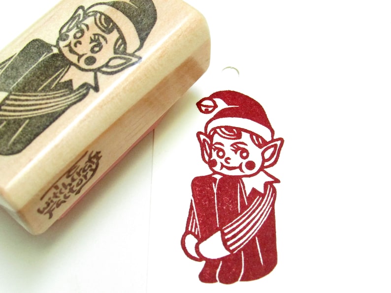 May include: A wooden stamp featuring a carved image of a smiling elf in a hat and collared shirt. Beside it is a red ink impression of the elf. The stamp has the text "witchcraft factory" on the side.