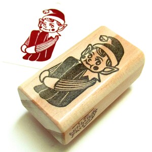 May include: A wooden stamp with a cartoon elf design. The elf is in a red outfit and hat with a black outline. A stamped image of the elf is visible on a white surface. The stamp has the text "with love, friends" on the side.