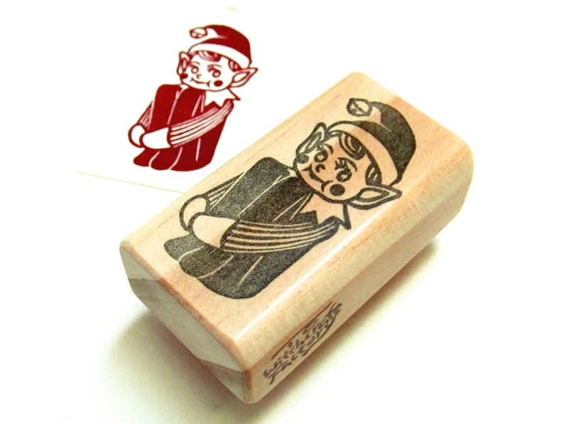 May include: A wooden stamp with a cartoon elf design. The elf is in a red outfit and hat with a black outline. A stamped image of the elf is visible on a white surface. The stamp has the text "with love, friends" on the side.