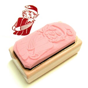 May include: A pink rubber stamp with a Christmas elf design, set on a light wood block. The stamp shows a detailed relief of the elf in a red outfit and hat. A red ink impression of the elf is visible on the left.