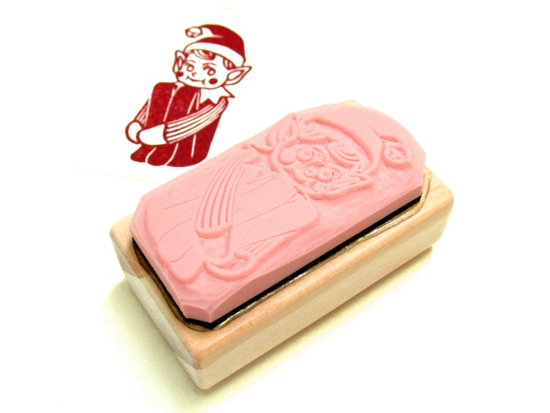 May include: A pink rubber stamp with a Christmas elf design, set on a light wood block. The stamp shows a detailed relief of the elf in a red outfit and hat. A red ink impression of the elf is visible on the left.