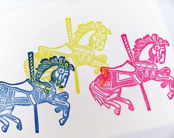May include: Three linocut prints of carousel horses in blue, yellow, and pink. Each horse features a decorative saddle and pole. The prints are set against a white background, with the horses overlapping.