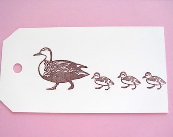 May include: A white tag featuring a brown ink illustration of a duck and three ducklings. The duck family is walking in a row. The tag has a hole punched in the upper left corner.