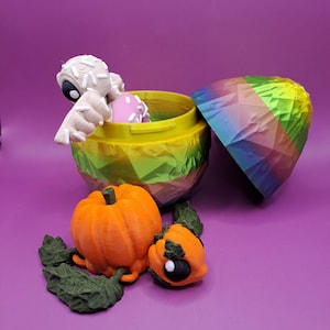 Puede incluir: Un recipiente impreso en 3D con una tapa de colores arcoíris y una base amarilla, que contiene una pequeña figura y un objeto rosa. Dos calabazas naranjas y hojas verdes están frente al recipiente. El fondo es morado.
