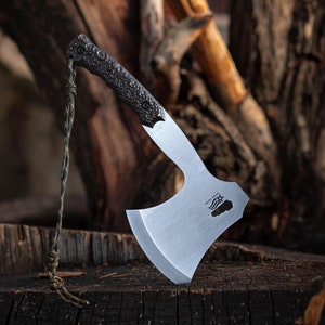 May include: A small, silver-colored axe with a black handle. The axe is sitting on a wooden surface.