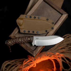 以下が含まれることがあります： 茶色の革製の鞘に、茶色の木製の柄と銀色の刃を持つナイフが入っています。ナイフは、茶色の革製の鞘が付いた木製の箱の上に置かれています。