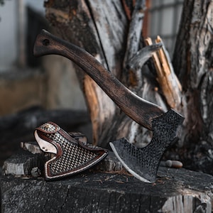 May include: Axe with a dark wooden handle and a textured, dark metal head. The axe rests on a tree stump next to a leather sheath with a woven pattern and brown trim. The wood has a natural, aged appearance.