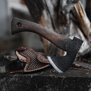 May include: A dark-handled axe with a textured black head and a leather sheath. The axe has a wooden handle and a sharp, silver blade. The sheath is brown and tan with a woven design.