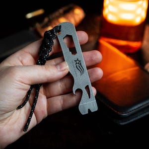 May include: A silver multi-tool with a bottle opener, star logo, and other functions. It has a black and white lanyard attached. The tool is held in a hand, with a blurred background.