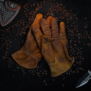 Puede incluir: Un par de guantes de trabajo de cuero marrón con costuras visibles y un acabado envejecido. Los guantes descansan sobre una superficie oscura salpicada de granos de café, con una funda de cuero y un cuchillo en el fondo.