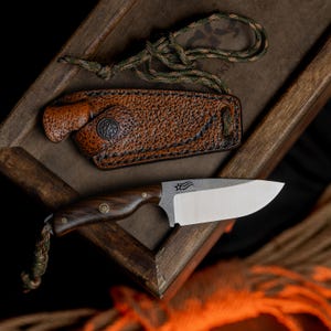 May include: A small, fixed blade knife with a brown wooden handle and a stainless steel blade. The blade has a star and stripes design etched into it. The knife is in a brown leather sheath with a green and brown paracord lanyard.