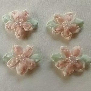 May include: Four small, fabric flower embellishments. Each flower has soft pink petals and a light green leaf base. The center of each flower has a cluster of small, clear beads. These decorative flowers are ideal for craft projects.