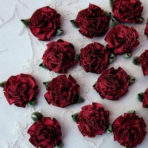 May include: A collection of small, dark red ribbon roses, each featuring tightly gathered fabric petals and a small green leaf accent at the base. These decorative floral embellishments are arranged in a scattered pattern.