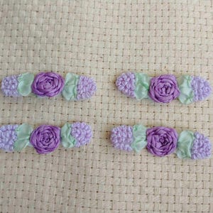 May include: Four handmade floral hair clips. Each clip features a central purple rose, flanked by light green leaves and lavender-colored, textured accents. The clips are arranged on a beige, woven background.