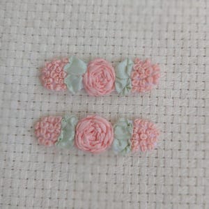 May include: Two handmade hair clips with a floral design. Each clip features a pink rose in the center, flanked by light green leaves and textured pink accents. The clips are set against a textured, off-white background.