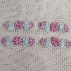May include: Four handmade floral embellishments. Each features a central pink rose, flanked by light green leaves and textured pink accents. The items are arranged on a textured, off-white fabric background.