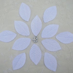 May include: Twelve white fabric leaves arranged in a circle on a white lace background. A silver rhinestone embellishment is in the centre of the circle.
