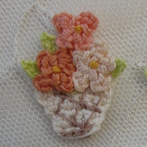 May include: A small embroidered flower arrangement in a basket. The flowers are pink, peach, and white. The basket is beige.