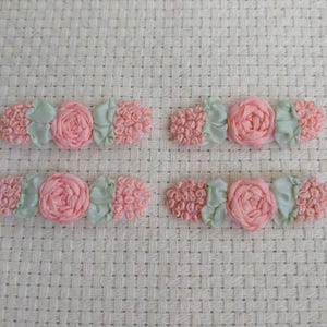 May include: Six handmade floral embellishments. Each features a pink rose, two clusters of pink textured fabric, and two light green fabric leaves. The items are arranged on a white woven background.