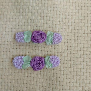 May include: Two decorative hair clips featuring a central purple rose surrounded by lavender-colored accents and light green leaves. The clips are set against a beige woven background, showcasing a floral design.