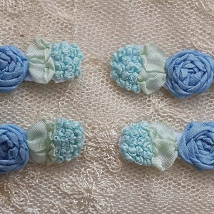 May include: Four handmade hair clips with a floral design. Each clip features a light blue rose in the center, flanked by light green and light blue fabric accents. The clips are arranged on a cream-colored, lace-like surface.