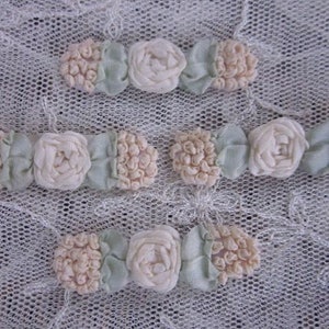May include: Three rows of delicate white and green fabric flowers, each row with a central white flower and two smaller white flowers on either side. The flowers are arranged on a white lace background.