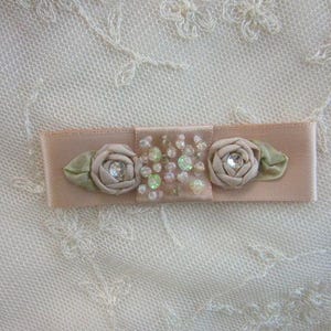 May include: A blush-colored ribbon hair clip with fabric roses and sparkling embellishments. The clip features two fabric roses with a rhinestone center and green leaves. The center section is adorned with clear and green rhinestones.