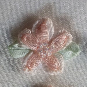 May include: A close-up of a delicate, handmade fabric flower. The flower has five petals in a soft peach color, with a cluster of small, clear beads at the center. Two light green leaves are positioned beneath the petals. The flower is likely used for crafting or embellishment.