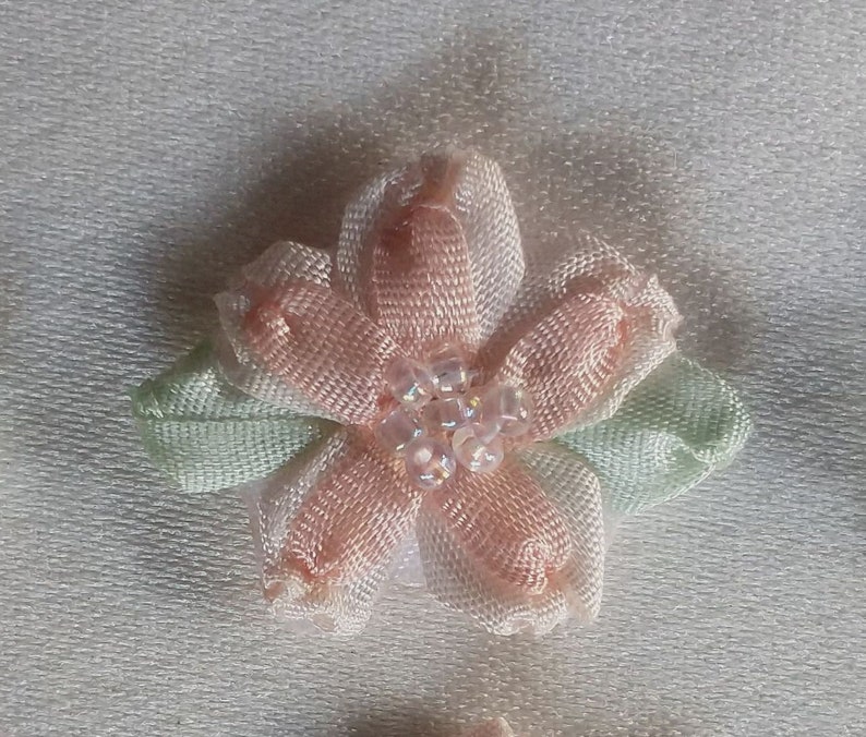 May include: A close-up of a delicate, handmade fabric flower. The flower has five petals in a soft peach color, with a cluster of small, clear beads at the center. Two light green leaves are positioned beneath the petals. The flower is likely used for crafting or embellishment.