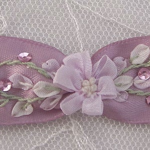 May include: A pink satin ribbon bow with a delicate floral design embroidered in white and green. The bow is decorated with small, iridescent sequins.