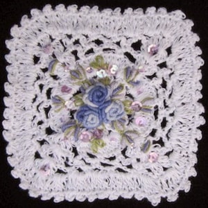 May include: White crocheted square with a floral design in the center. The flowers are blue and purple with pink and green accents. The square is decorated with iridescent sequins.
