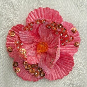 May include: A pink velvet flower with a ruffled edge, decorated with gold sequins. The flower has a center made of a clear orange gem.