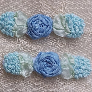 May include: Two hair barrettes with a floral design. Each barrette features a central light blue rose flanked by light blue and pale green fabric accents. The barrettes are set against a textured, off-white background.