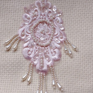May include: A delicate pink lace applique with pearl bead embellishments. The lace features a floral design with a central circle of pearls. The applique has a fringe of pearls hanging from the bottom.