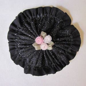 May include: A black fabric rosette with a ruffled edge and a center decoration of three pink fabric roses with green leaves.