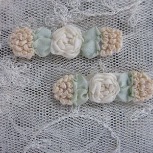 May include: Two small hair clips with floral designs. Each clip features a central white fabric rose, flanked by light green fabric and textured beige fabric. The clips rest on a white lace background.
