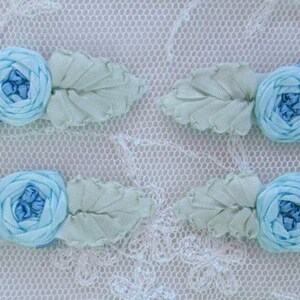 May include: Four handmade floral hair accessories. Each features a light blue fabric rose with matching leaves. The leaves are a pale green color. The background is a white lace fabric.
