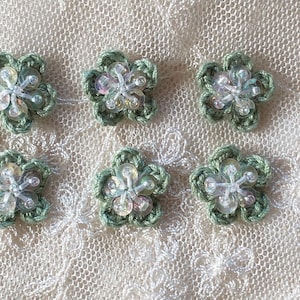 May include: Six small, handmade crochet flowers in a light green shade, each with clear, iridescent bead centres. They are set on white, textured lace fabric, giving a delicate, feminine look. Suitable for embellishments or craft projects.