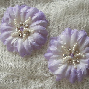 May include: Two lavender fabric flowers with white centers and pearl and rhinestone embellishments.