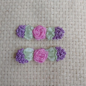 May include: Two decorative hair clips. Each clip features a pink rose in the center, flanked by light green and purple floral accents. The clips are set against a textured, off-white background.