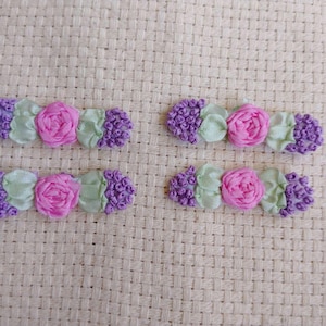 May include: Four handmade floral embellishments. Each features a pink rose, light green leaves, and clusters of purple buds. The floral elements are arranged on a light beige woven background, creating a delicate and textured appearance.