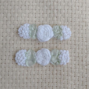 May include: Two white fabric hair clips. Each clip features a central white rose, flanked by white pom-poms and light green ribbon accents. The clips are set against a textured, off-white background.