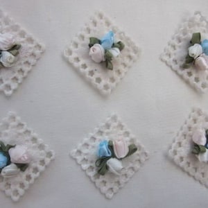 May include: Six white crocheted squares with a cluster of three satin ribbon flowers in the center of each square. The flowers are in shades of blue and pink.