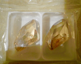 May include: Two faceted, amber-colored crystal pendants are displayed in a clear plastic package. The crystals have a pointed shape with multiple cut surfaces, reflecting light. The pendants are set in a white plastic tray.